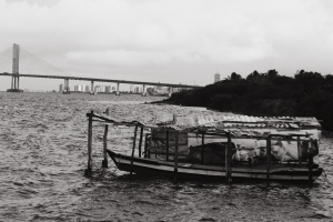 A obra de Eric de Sousa Estevao se chama Prainha Resiste e é um conjunto de quatro fotografias de uma comunidade tradicional pesqueira de Aracaju, localizada à margem do Rio Sergipe. A primeira fotografia mostra um barco de madeira entre duas palafitas. Palafitas é um tipo de casa de madeira que é erguida com estacas em locais alagados ou em margens de rios. Ao fundo, aparece a outra margem do rio na linha do horizonte. A segunda fotografia mostra uma palafita a lateral de uma palafita no lado direito da imagem. No lado esquerdo há quatro barcos sobre o rio, ao fundo, aparece a outra margem do rio na linha do horizonte. Na terceira fotografia há um barco visto de lado, de frente para a esquerda, coberto por um telhado de placas sustentado por estacas de madeira. Ao fundo no lado direito vemos a ponta do território cheio de vegetação. Ao fundo no lado esquerdo numa distância longa, há uma ponte e vários prédios na linha do horizonte. Na quarta fotografia há uma pequena palafita de parede branca, com um portão de madeira, entre duas casas. Ao fundo, atrás da casa branca, aparece a outra margem do rio na linha do horizonte. 