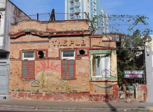 A obra de Djonathan Freitas se chama Veracidade Arruinada é um conjunto de três fotografias de fachadas casas em estado avançado de deteriorização. A primeira fotografia mostra a fachada de uma casa ao centro de parede cinza, com um portão de grade de cor preta ao lado direito. Há quatro janelas, duas delas estão fechadas, uma está aberta e uma delas, a janela mais à direita, possui uma grade de cor preta e uma placa preta pendurada com a frase "o resto é só de fachada" escrita com letras de cor branca e vermelha. Há algumas pixações da parede, uma delas mais à direita e na parte inferior com as palavras fora Temer escritas em preto. A segunda fotografia mostra uma casa com a fachada de cor bege, com tijolos expostos. A parede tem três janelas, sendo duas delas com molduras vermelhas e fechadas e uma janela aberta à direita de moldura branca, com plantas na parte superior. À direita desta janela, há um portão com uma placa de cor preta pendurada com as palavras veracidade arruinada escritas em letras brancas e rosas. O portão tem uma grade preta que vai até a parte de cima da fachada da casa. Em cima da janela do meio, há escritas as palavras The Pub, com letras na mesma cor da parede. A terceira fotografia mostra um poste de fios elétricos no lado esquerdo da imagem. Há um muro de cor cinza escuro, com pixações e tinta antiga descascando com uma placa preta ao centro com as palavras lembre-se escritas com letras brancas e rosas. Acima do muro há uma grade de fios elétricos bem finos. Atrás do muro há uma casa branca bem antiga, com janelas bem altas e sem molduras, apenas com um espaço aberto. 