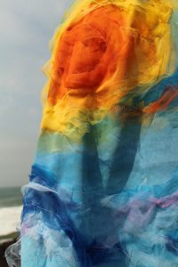 A obra de Ana Carolina Hiera Camargo se chama Costurando a Paisagem e é um conjunto de cinco fotografias de uma mulher de cabelos castanhos e ondulados em uma praia. A mulher está sobre um chão de pedras, um pouco distante do mar. Na primeira fotografia, a mulher está de costas, descalça, usa uma calça branca e um casaco azul claro, com detalhes verdes na parte debaixo, detalhes azuis e roxos ao longo da roupa e há um sol amarelo e laranja feito com um tecido fixado no meio das costas. A segunda fotografia mostra de perto o sol das costas do casaco e nesta imagem é possível notar que os detalhes em azul escuro também são feitos a partir de um tecido fixado sobre o casaco. Na terceira fotografia, a mulher está de perfil, com a cabeça inclinada para a esquerda, com as pedras e o mar ao fundo. Na quarta fotografia, a mulher é mostrada mais de perto do que na primeira fotografia. Ela está de costas, com o braço direito dobrado, a mão na cabeça, com o pescoço virado para trás, sendo possível ver seus óculos. A quinta fotografia mostra a superfície do casaco em detalhe, com o mar ao fundo. 