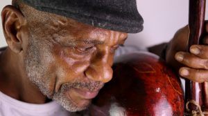 A obra de Daniel Souza do Nascimento se chama Ginga capoeirista: Conexão entre capoeira e fotografia e é composta por três imagens retangulares horizontais. Na primeira há um homem negro de boina preta e camisa branca tocando um berimbau. A fotografia mostra o rosto do homem, bem próximo à cabaça com textura vermelha do berimbau, a mão esquerda do homem segura o arco do instrumento. Na segunda fotografia há um homem negro descalço usando calça e camisa brancas visto de cima, com a mão no chão e o braço direito dobrado, que por sua vez apoia suas duas pernas dobradas. Na terceira fotografia há um homem negro e uma mulher negra de calças e camisas brancas. O homem está ao lado esquerdo, de perfil, virado para a direita, usa óculos, cordel verde amarrado na cintura e está em pé com o corpo curvado para trás. A mulher está do lado direito, com as mãos no chão, com a perna esquerda esticada com pé no chão, perna direita esticada para cima apontada na direção do homem como se aplicasse um golpe. A mulher usa cordel azul amarrado na cintura e cabelo preso. Os dois estão na frente de uma casa feita de pedaços de madeira. 